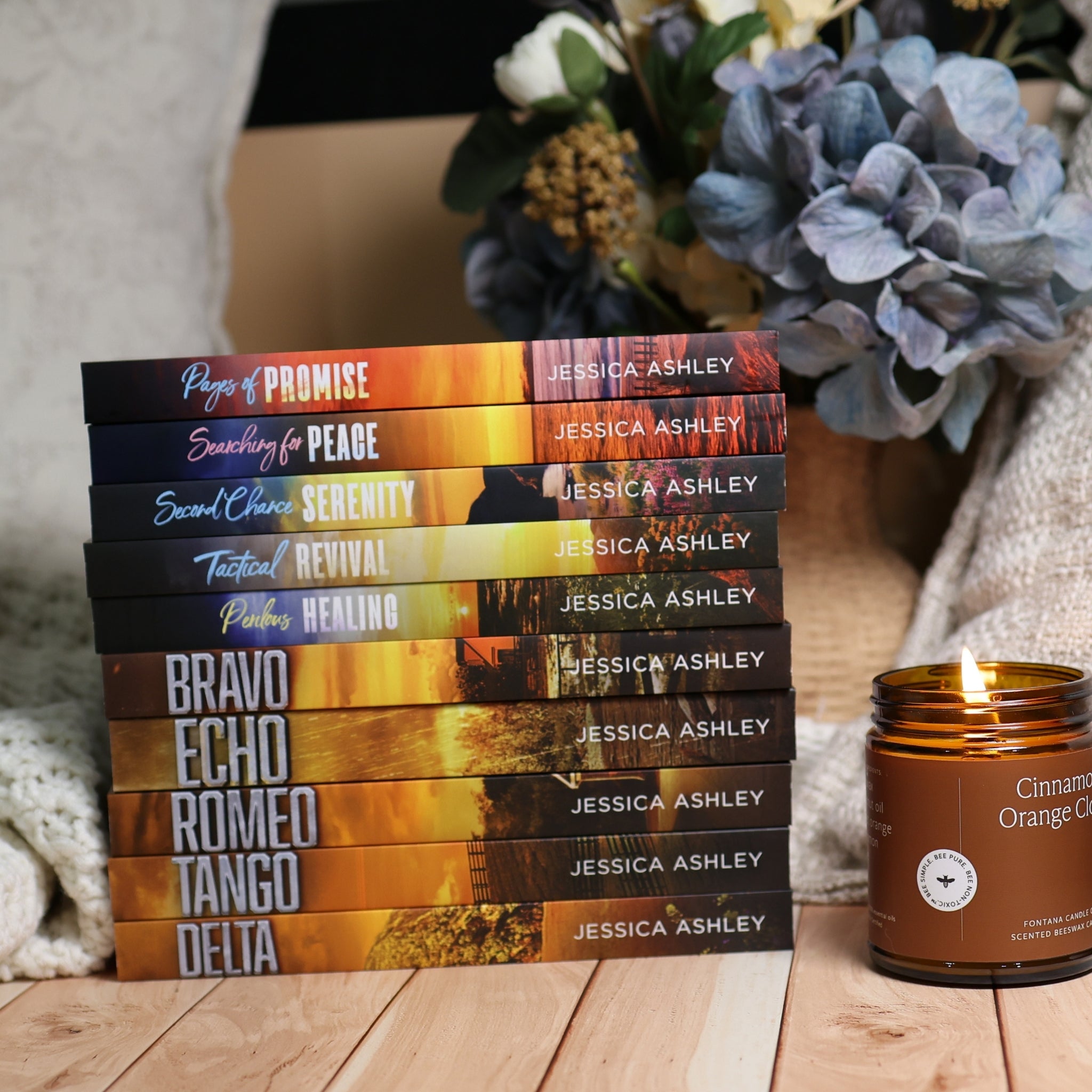 A stack of ten books by Jessica Ashley with dark, dramatic romance novel covers is arranged on a wooden surface next to a lit amber candle. The background features a cozy neutral wall, a soft knit blanket, and a decorative floral arrangement.
