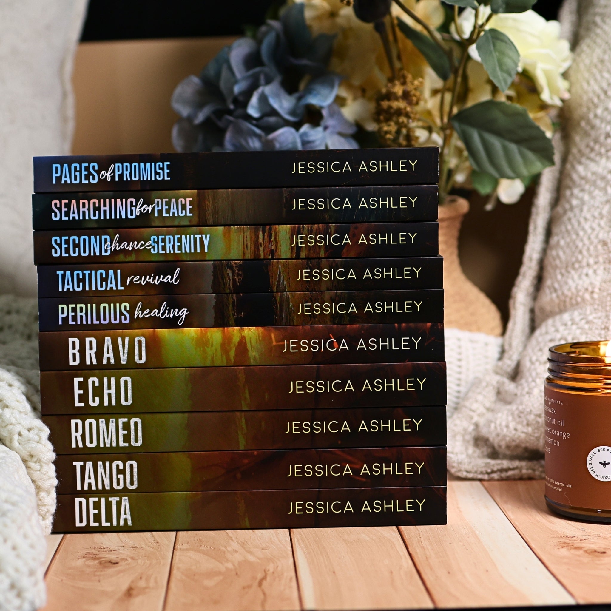 A stack of ten books by Jessica Ashley, titled Pages of Promise to Tango Delta, arranged on a wooden surface with a cozy knit blanket, a candle, and a vase of flowers in the background.