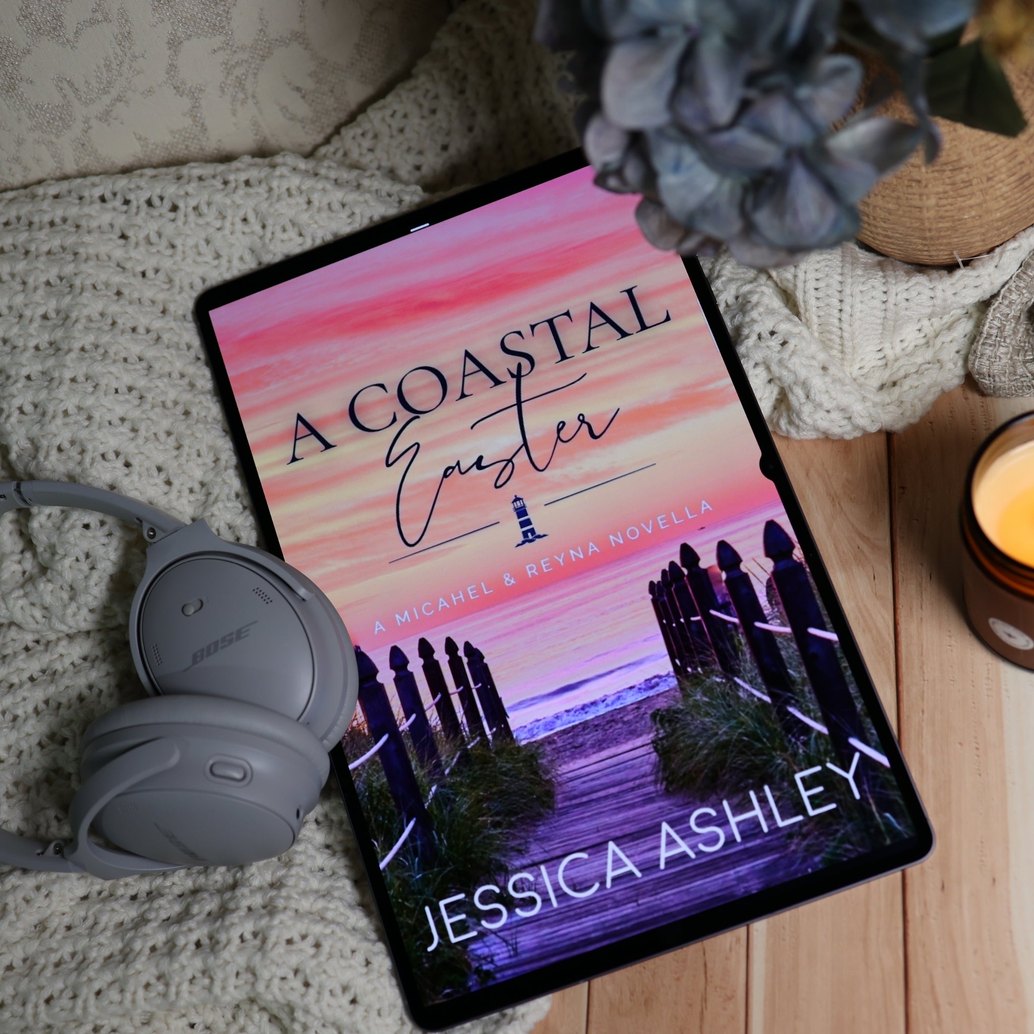 A tablet lying on a cream knit blanket on a wooden surface, displaying a book cover titled "A Coastal Easter" by Jessica Ashley. Gray Bose headphones rest beside the tablet, with a blue flower, woven decor, and a lit candle in the background.