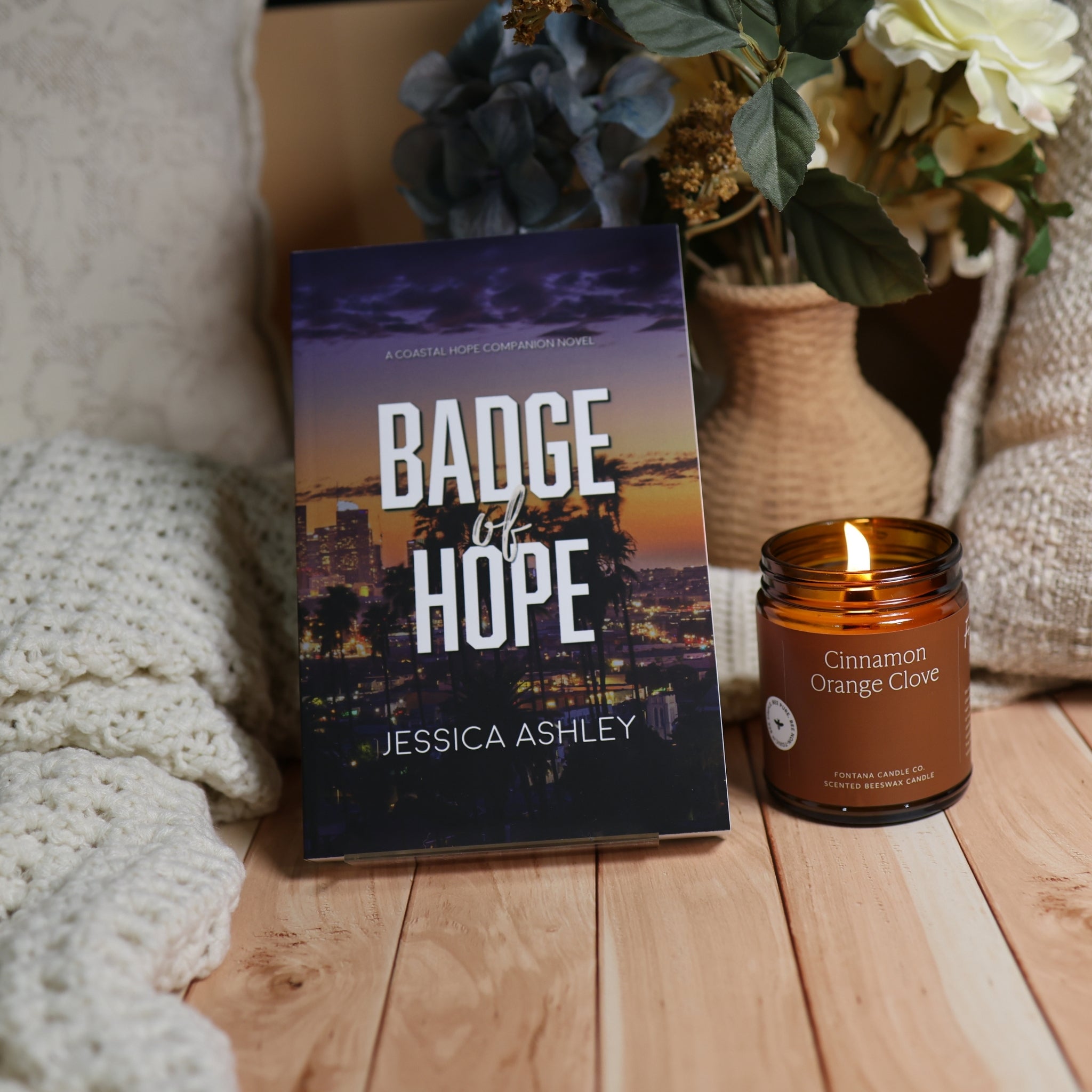 A book titled 'Badge of Hope' by Jessica Ashley stands upright on a wooden surface next to a lit amber glass candle labeled 'Cinnamon Orange Clove.' The scene includes a knitted blanket, potted flowers, and a cozy home-like setting.