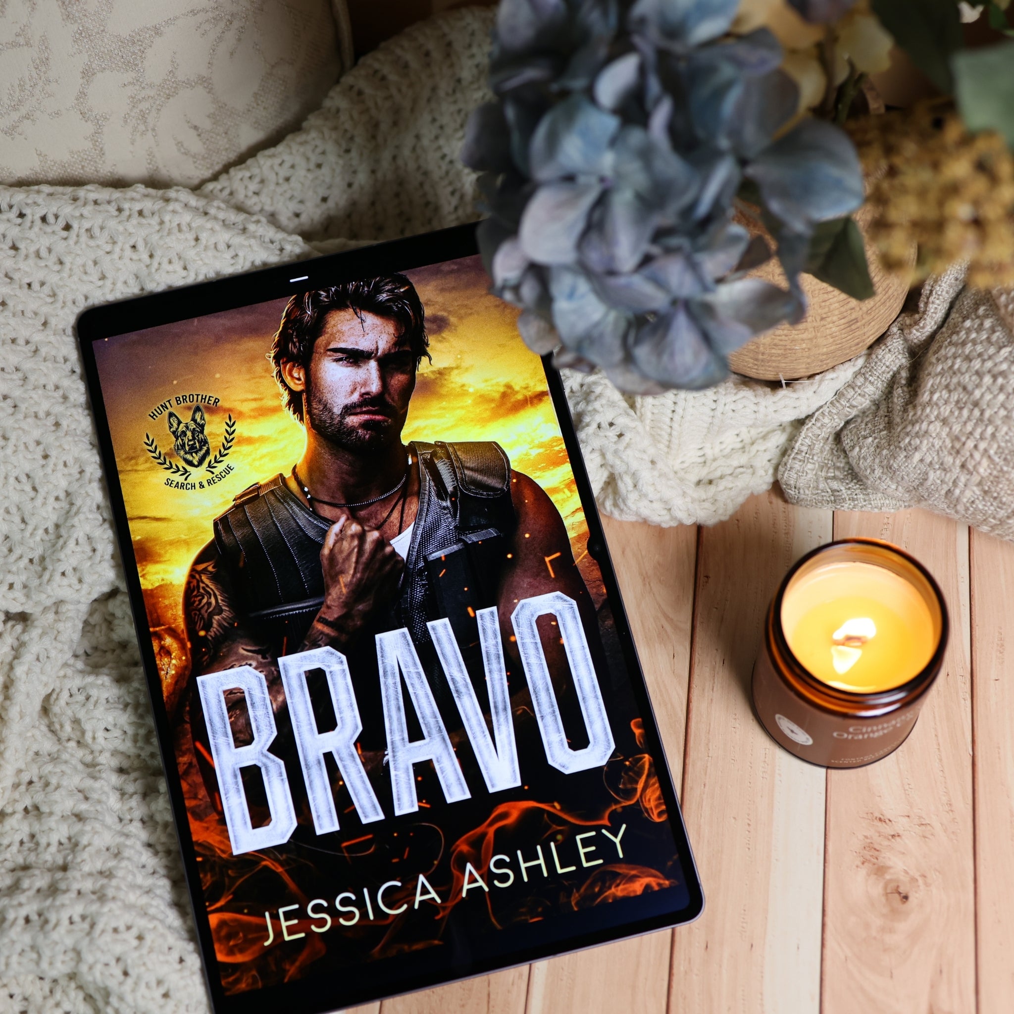 A tablet lying on a light wood surface, displaying a book cover for "Bravo" by Jessica Ashley. The scene is styled with a cream knit blanket, a blue hydrangea, and a lit amber candle.