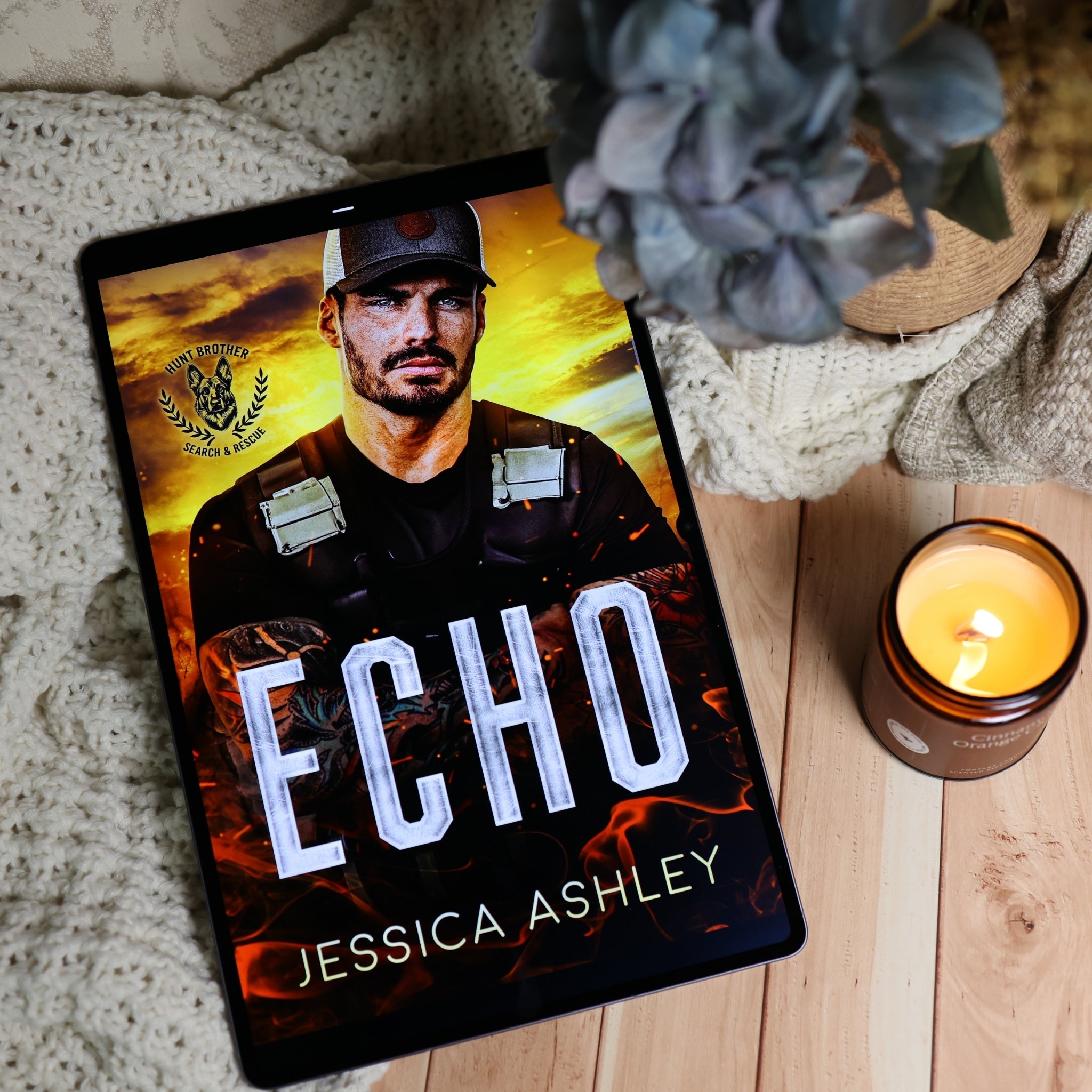 A tablet displaying the book cover of 'Echo' by Jessica Ashley, styled in a cozy flat-lay setting with a knitted blanket, a lit candle, and a potted plant on a light wood surface.