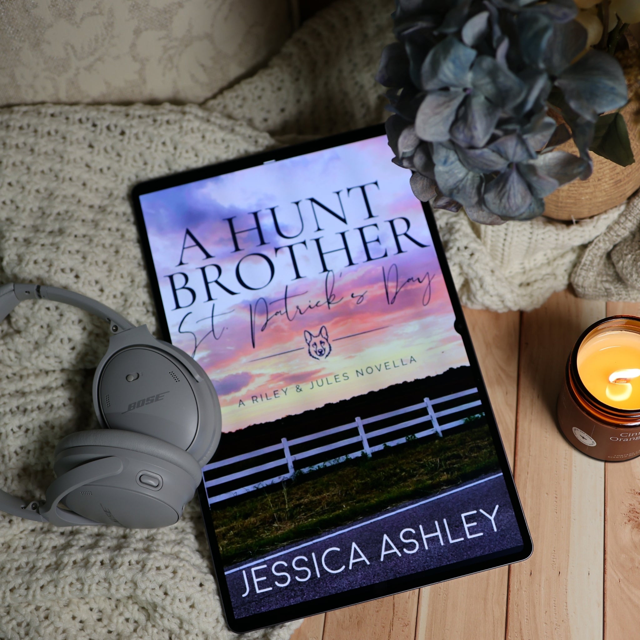 A tablet lying on a knitted blanket with a book cover for 'A Hunt Brother: St Patrick's Day' by Jessica Ashley, styled with gray Bose headphones, a blue hydrangea, and a lit candle on a wooden surface.