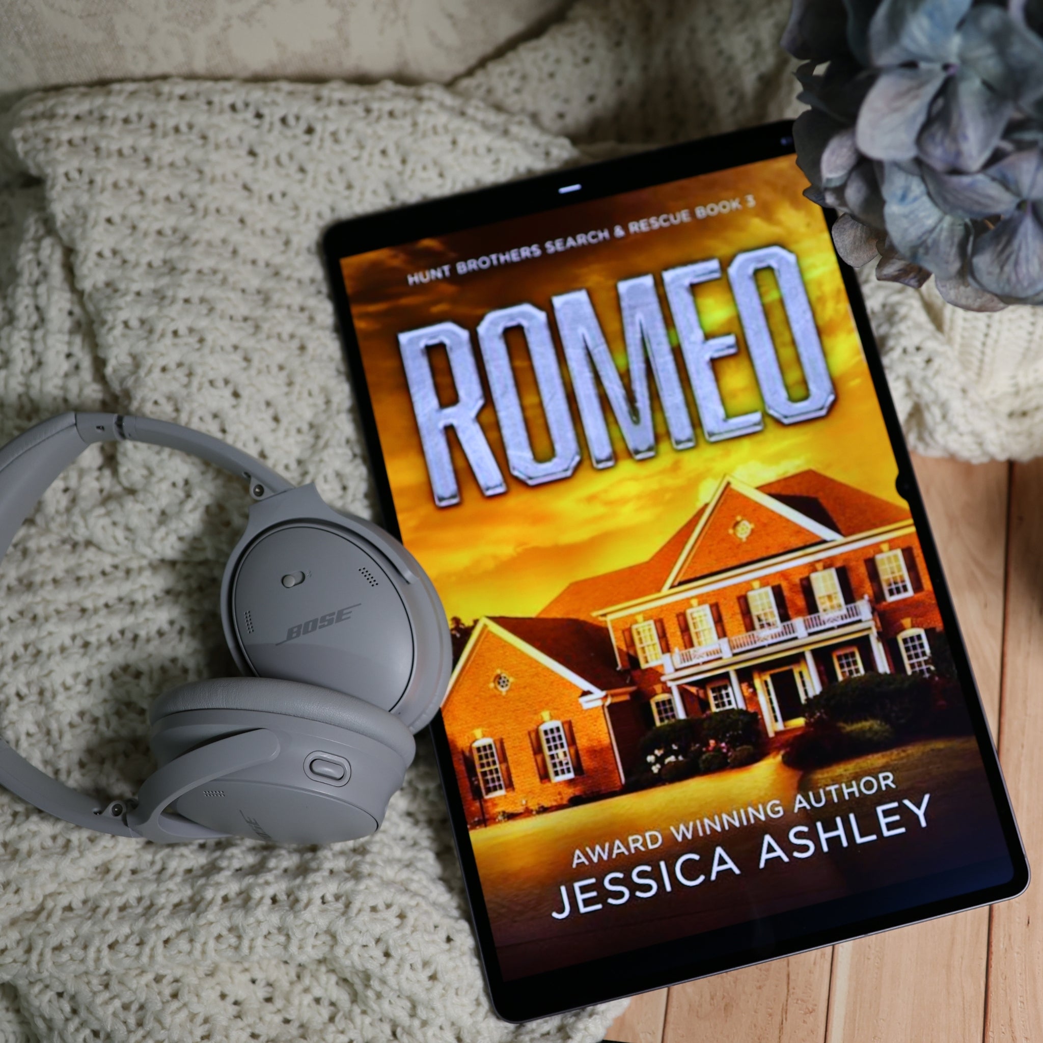 A tablet lying on a cream knit blanket with gray Bose headphones beside it and a dried flower in the corner. The screen shows a book cover titled 'ROMEO' by Jessica Ashley, with a house illustration and warm sunset tones.