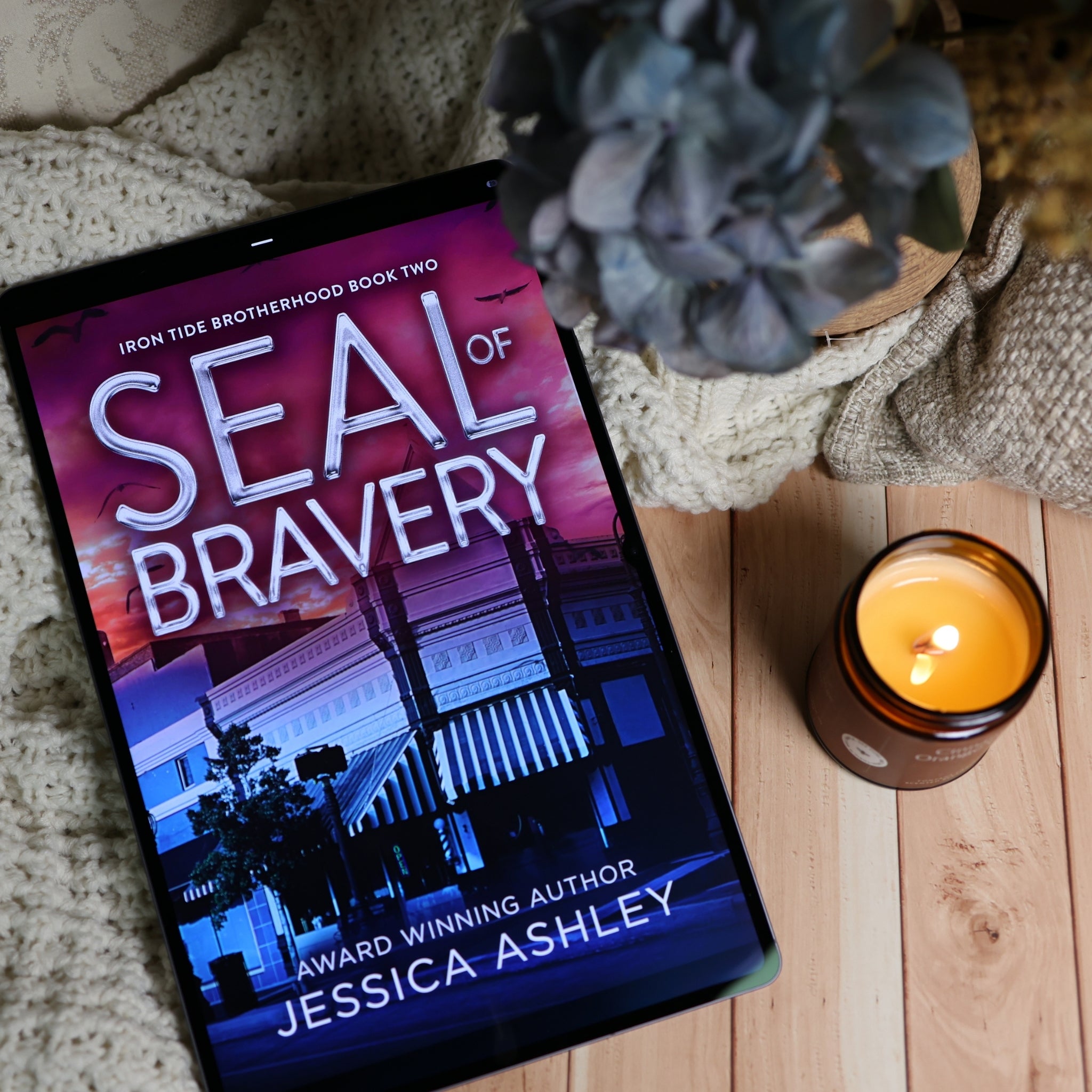 A tablet displaying the book cover of 'Seal of Bravery' by Jessica Ashley, styled on a wooden surface with a knitted blanket, a lit candle, and a potted plant.