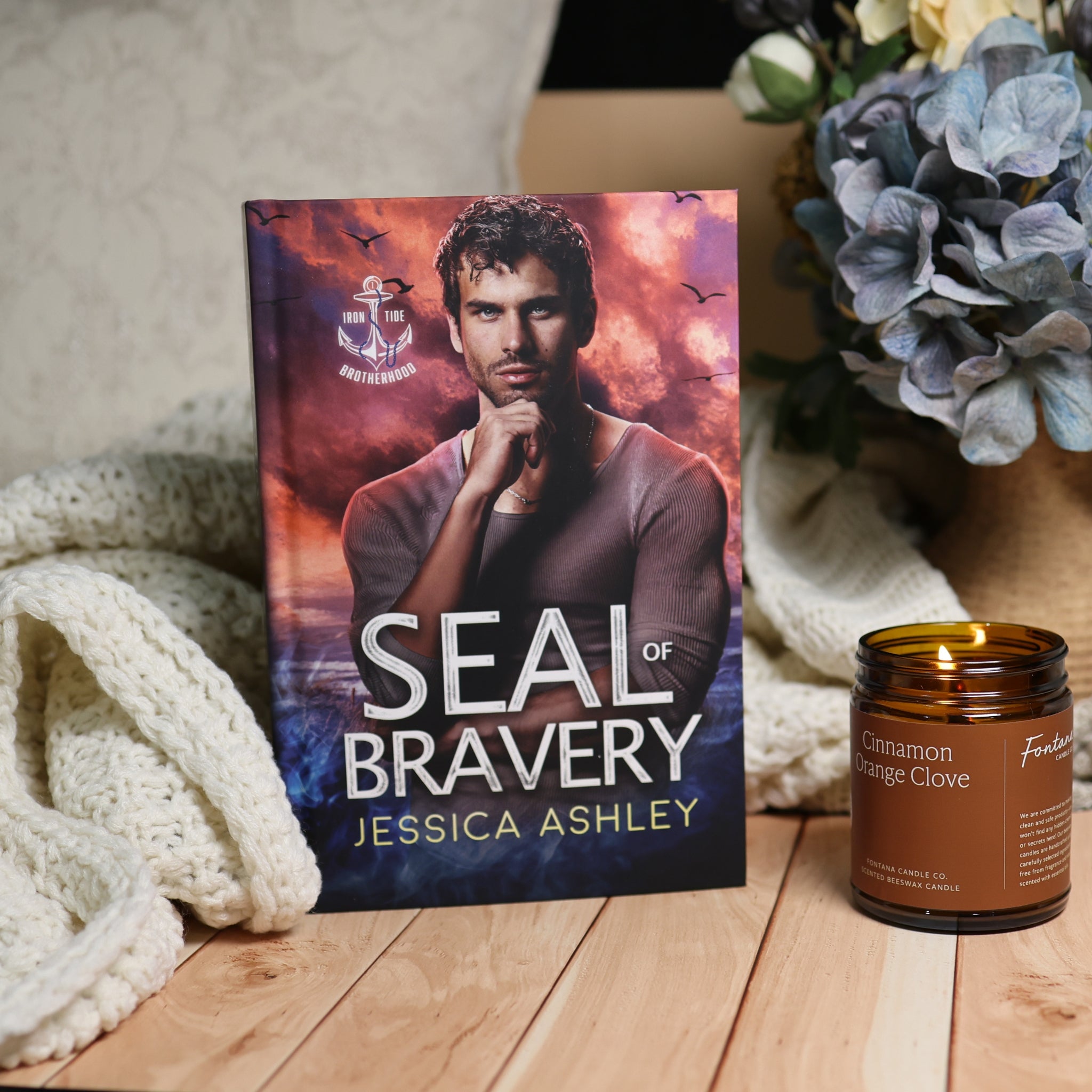 The book 'Seal of Bravery' by Jessica Ashley is displayed on a wooden table next to a lit candle and a cozy knit blanket, with a blurred background of flowers.