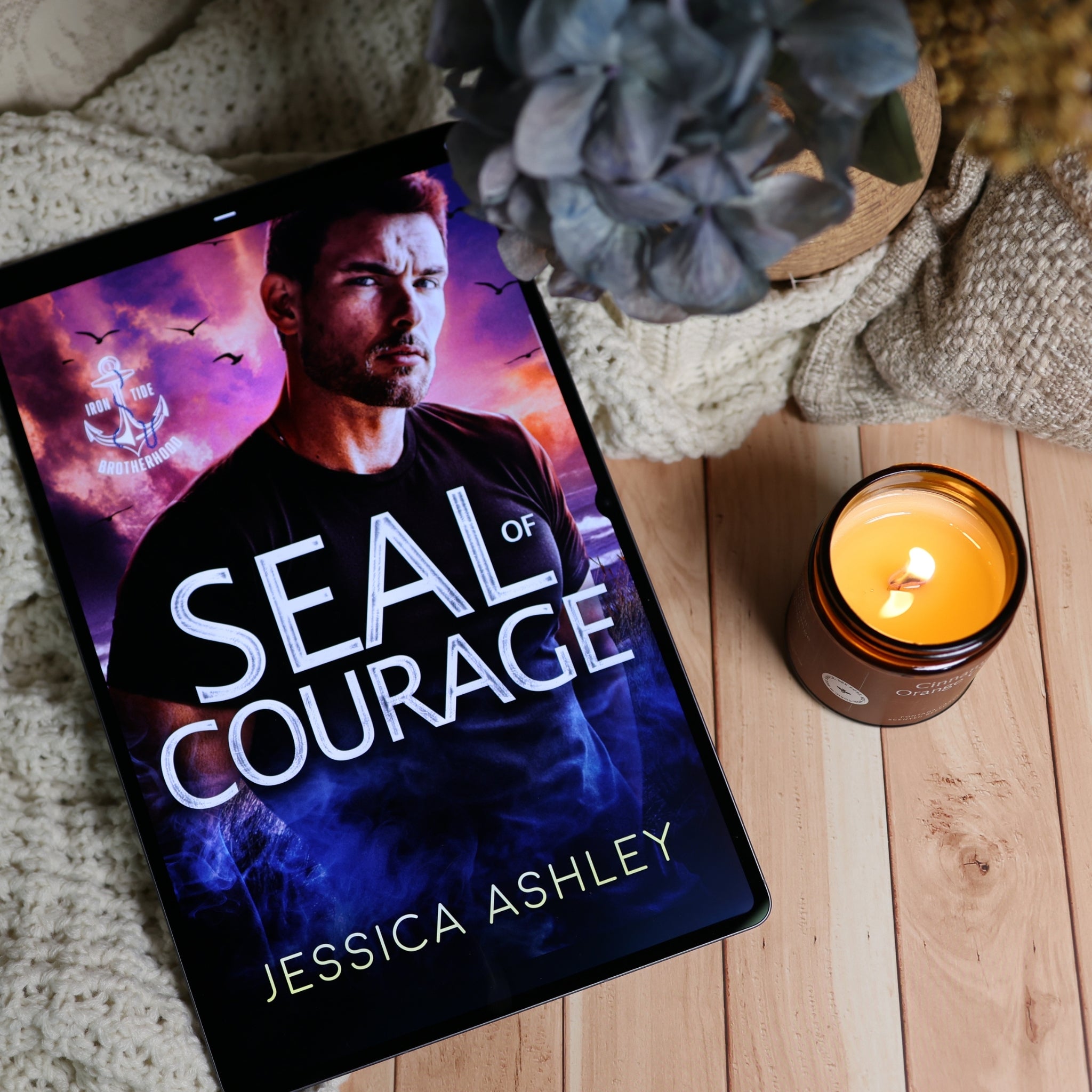 A tablet lying on a light wood surface displays the book cover for 'Seal of Courage' by Jessica Ashley. The scene is styled with a knitted blanket, a lit candle, dried flowers, and a blue-gray flower in the background.