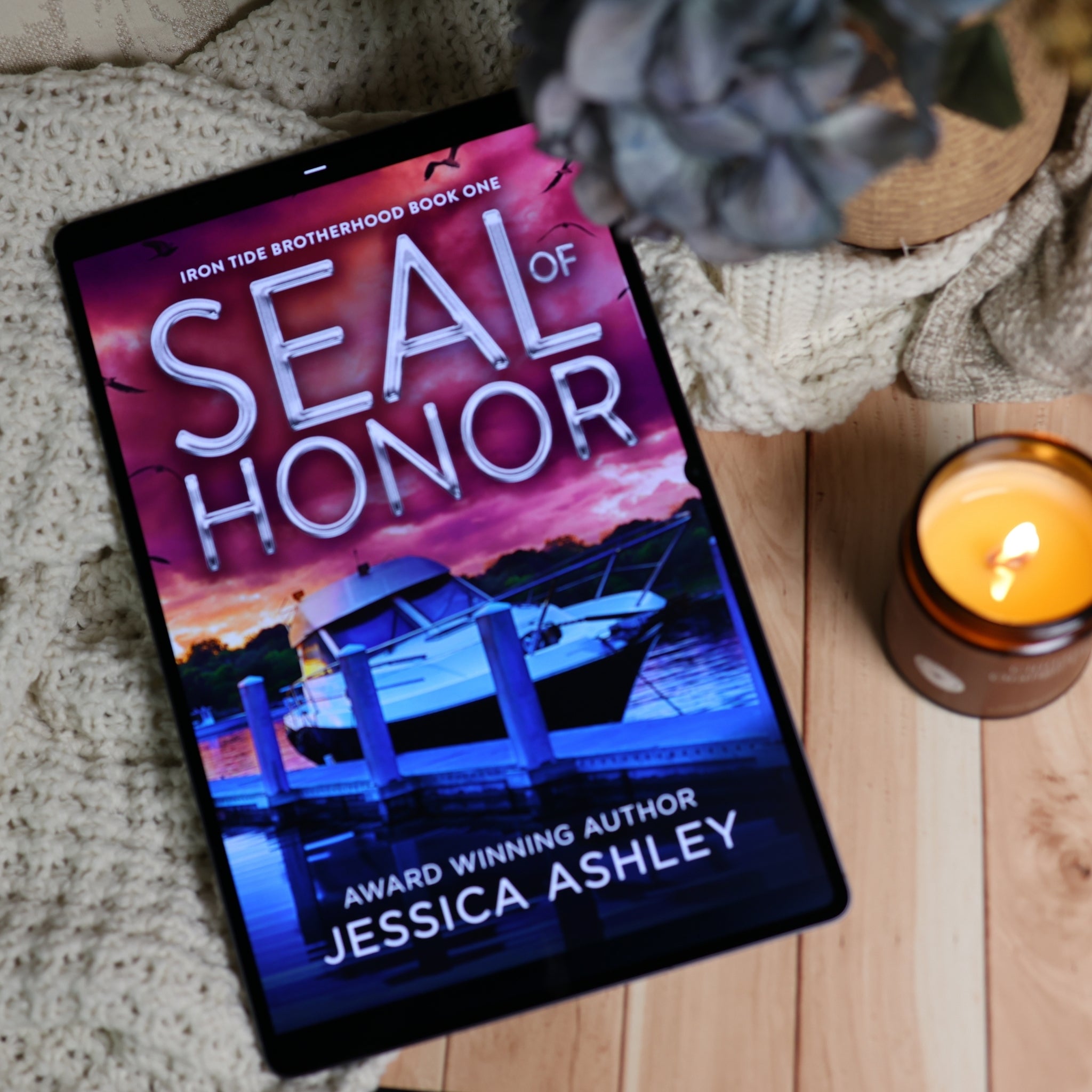 A tablet lying on a knitted blanket over a wooden surface, displaying the book cover for 'Seal of Honor' by Jessica Ashley. A lit candle and a potted plant are nearby in a cozy styled setting.