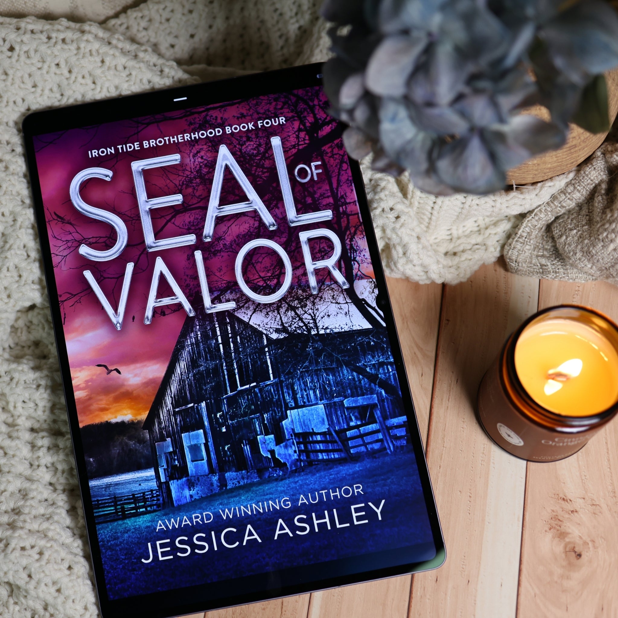 A tablet displaying the book cover of 'Seal of Valor' by Jessica Ashley, styled on a wooden surface with a knitted blanket, a lit candle, and a blue hydrangea flower.
