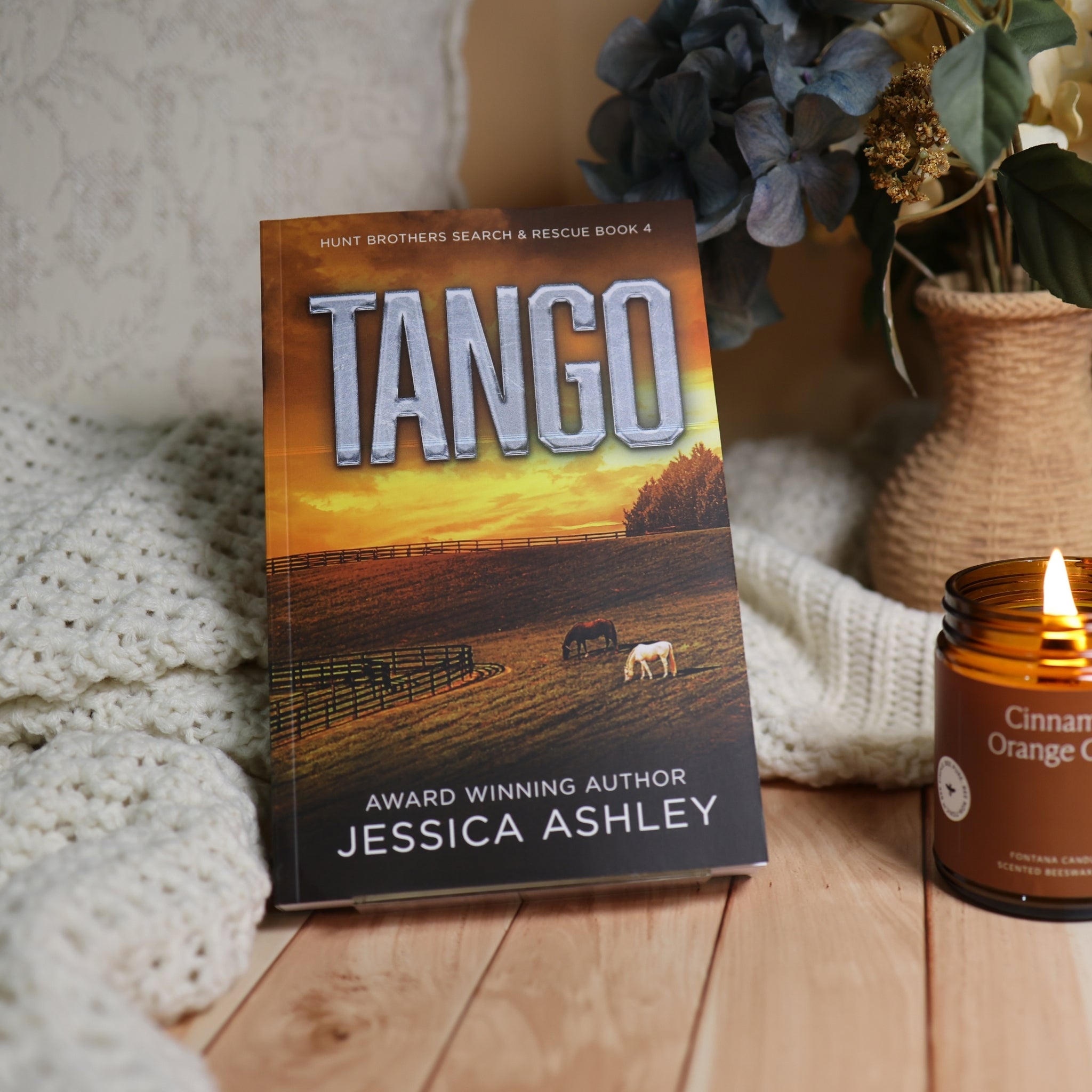 A book titled "Tango" by Jessica Ashley stands upright on a wooden surface, styled with a knitted cream blanket, a lit amber candle, and dried flowers in a woven vase.