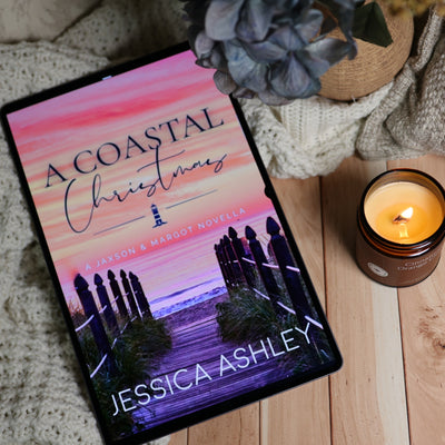 A tablet displaying the book cover of 'A Coastal Christmas' by Jessica Ashley, styled on a wooden surface with a knitted blanket, a lit candle, and a dried flower.