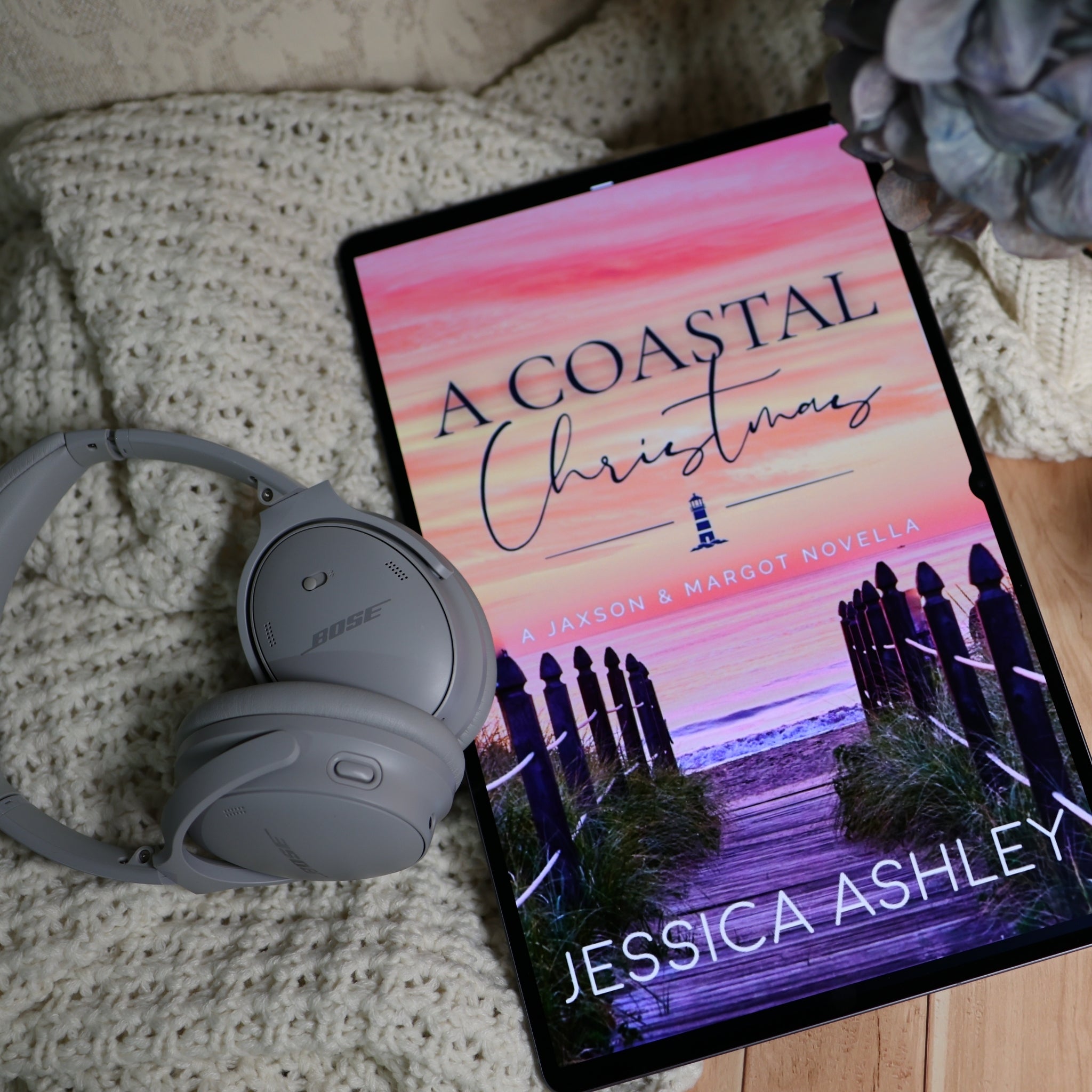 A tablet displaying the book cover of 'A Coastal Christmas' by Jessica Ashley, styled on a cream knit blanket with gray Bose headphones and a decorative flower.