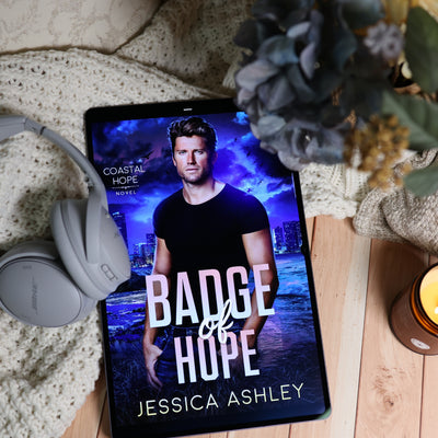A tablet lying on a wooden surface, displaying the book cover for 'Badge of Hope' by Jessica Ashley. The scene is styled with a cream knit blanket, gray over-ear headphones, dried flowers, and a lit candle.