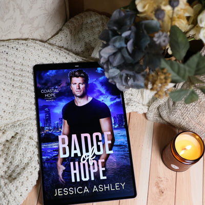 A tablet lying on a wooden surface, displaying the book cover for 'Badge of Hope' by Jessica Ashley. The scene is styled with a cream knit blanket, a lit amber candle, and dried flowers in the background.