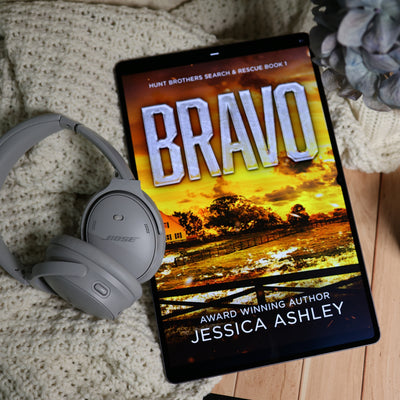 A tablet lying on a cream knit blanket with gray Bose headphones beside it. The screen displays the book cover for "Bravo" by Jessica Ashley, with a sunset farm scene and the title in large white letters.