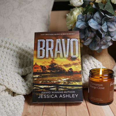 Book cover for 'Bravo' by Jessica Ashley displayed on a wooden surface next to a lit 'Cinnamon Orange Clove' candle, with a cozy white knit blanket and blue hydrangeas in the background.