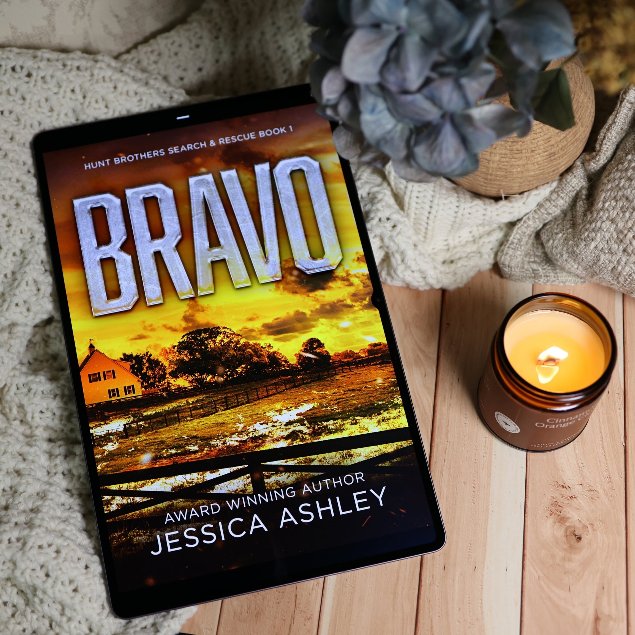 A tablet displaying the book cover of 'Bravo' by Jessica Ashley, styled on a wooden surface with a knitted blanket, a lit candle, and a potted plant.