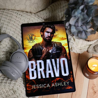 A tablet lying on a beige knit blanket and wood floor, displaying a book cover titled "BRAVO" by Jessica Ashley. Gray Bose headphones rest on the left, a blue hydrangea flower is on the upper right, and a lit amber candle is on the lower right.