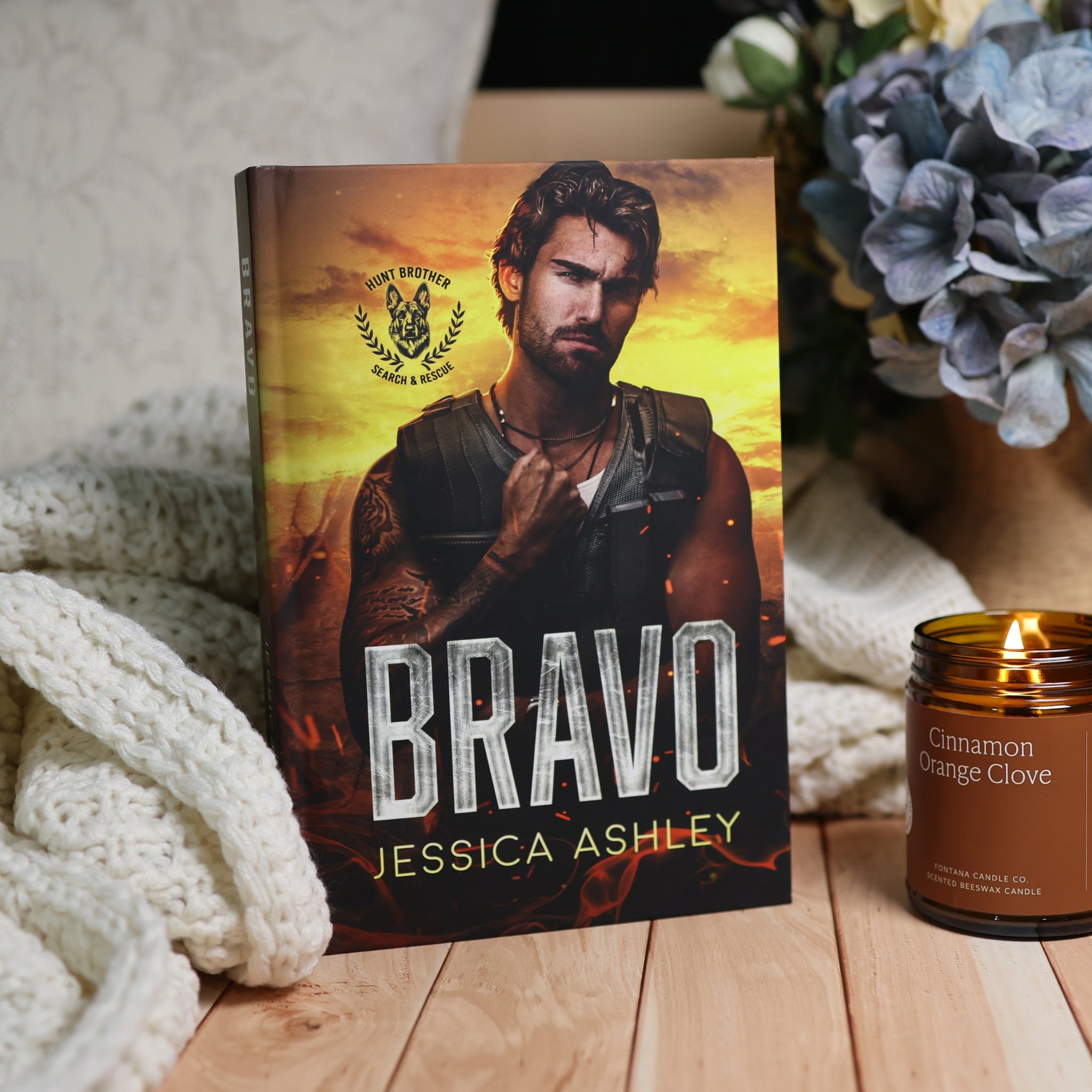 A hardcover book titled 'Bravo' by Jessica Ashley stands upright on a wooden surface, styled with a cream knit blanket, a lit amber candle, and blue flowers in the background.
