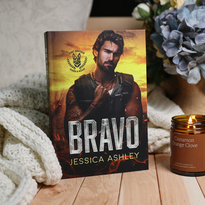 A hardcover book titled 'Bravo' by Jessica Ashley stands upright on a wooden surface, styled with a cream knit blanket, a lit amber candle, and blue flowers in the background.