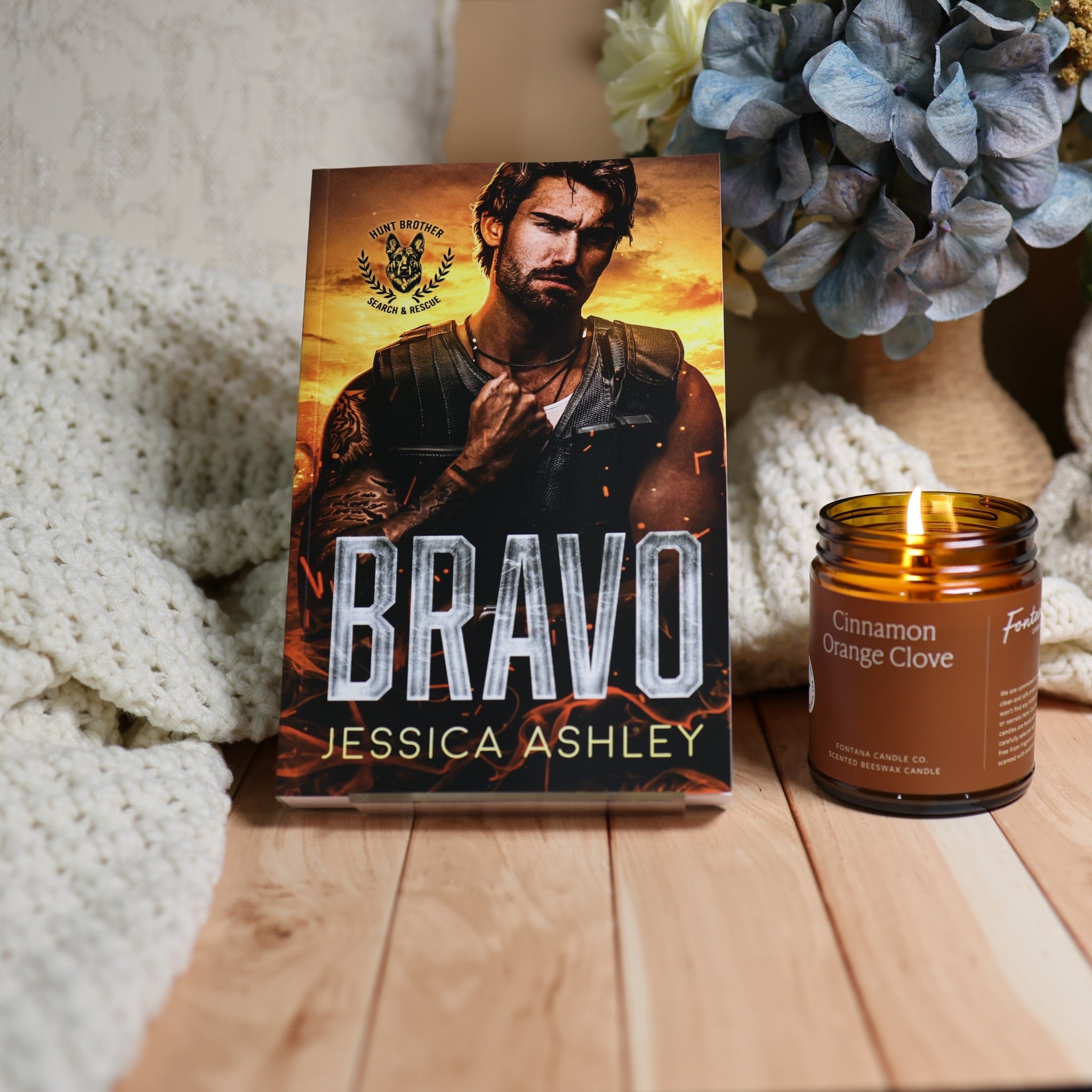 The book 'Bravo' by Jessica Ashley stands upright on a wooden surface next to a 'Cinnamon Orange' candle, styled with a cozy white knit blanket and blue hydrangeas in the background.