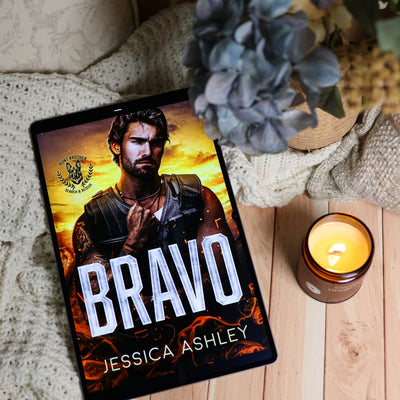 A tablet lying on a light wood surface, displaying a book cover for "Bravo" by Jessica Ashley. The scene is styled with a cream knit blanket, a blue hydrangea, and a lit amber candle.