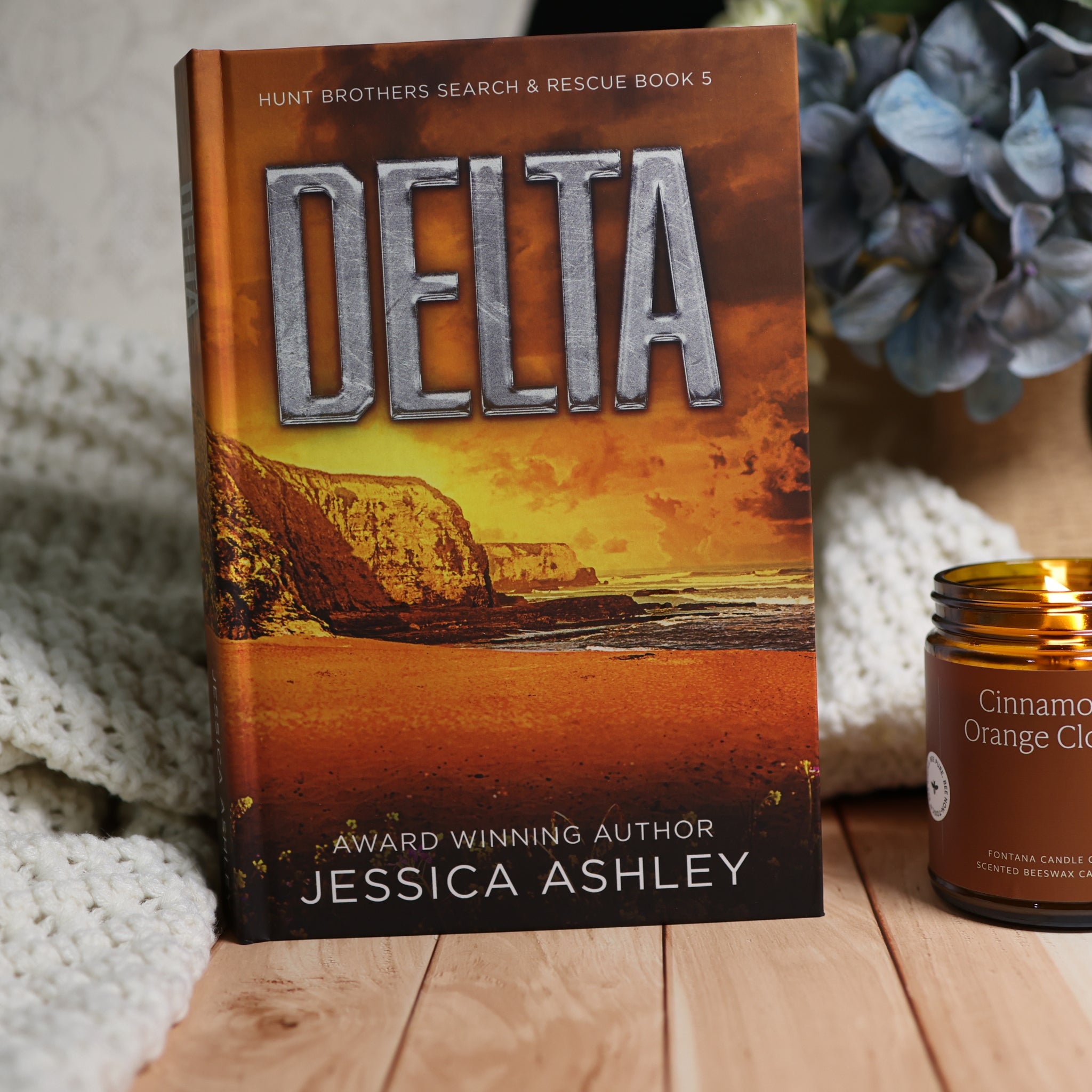 A hardcover book titled 'DELTA' by Jessica Ashley stands upright on a wooden surface, styled with a white knit blanket and a lit candle in the background.