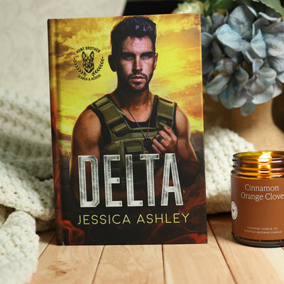 A hardcover book titled 'Delta' by Jessica Ashley stands upright on a wooden surface, styled with a knitted cream blanket on the left and a lit candle in an amber glass jar on the right. The book cover features a man in a military vest against a fiery yellow-orange background.