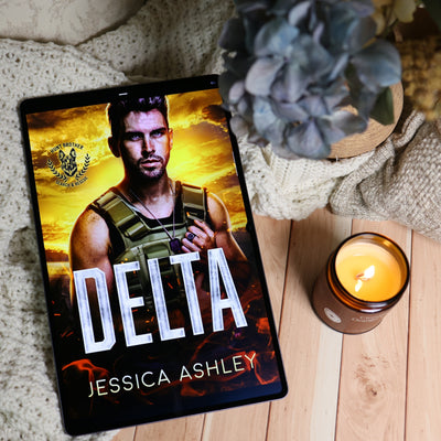 A tablet lying on a light wood surface, displaying a book cover titled "DELTA" by Jessica Ashley. The scene is styled with a knitted blanket, a blue-gray flower, and a lit candle.