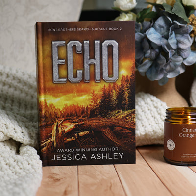 The book 'Echo' by Jessica Ashley stands upright on a wooden surface next to a 'Cinnamon Orange' candle, styled with a cozy white knit blanket and blue hydrangeas in the background.