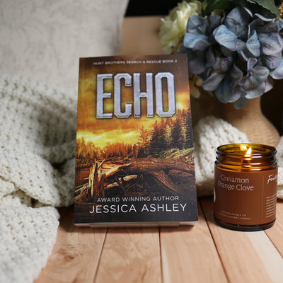 Book cover for 'Echo' by Jessica Ashley displayed upright on a wooden surface next to a lit amber candle jar labeled 'Cinnamon Orange Clove'. A knitted cream blanket and blue hydrangeas in a vase are softly blurred in the background.