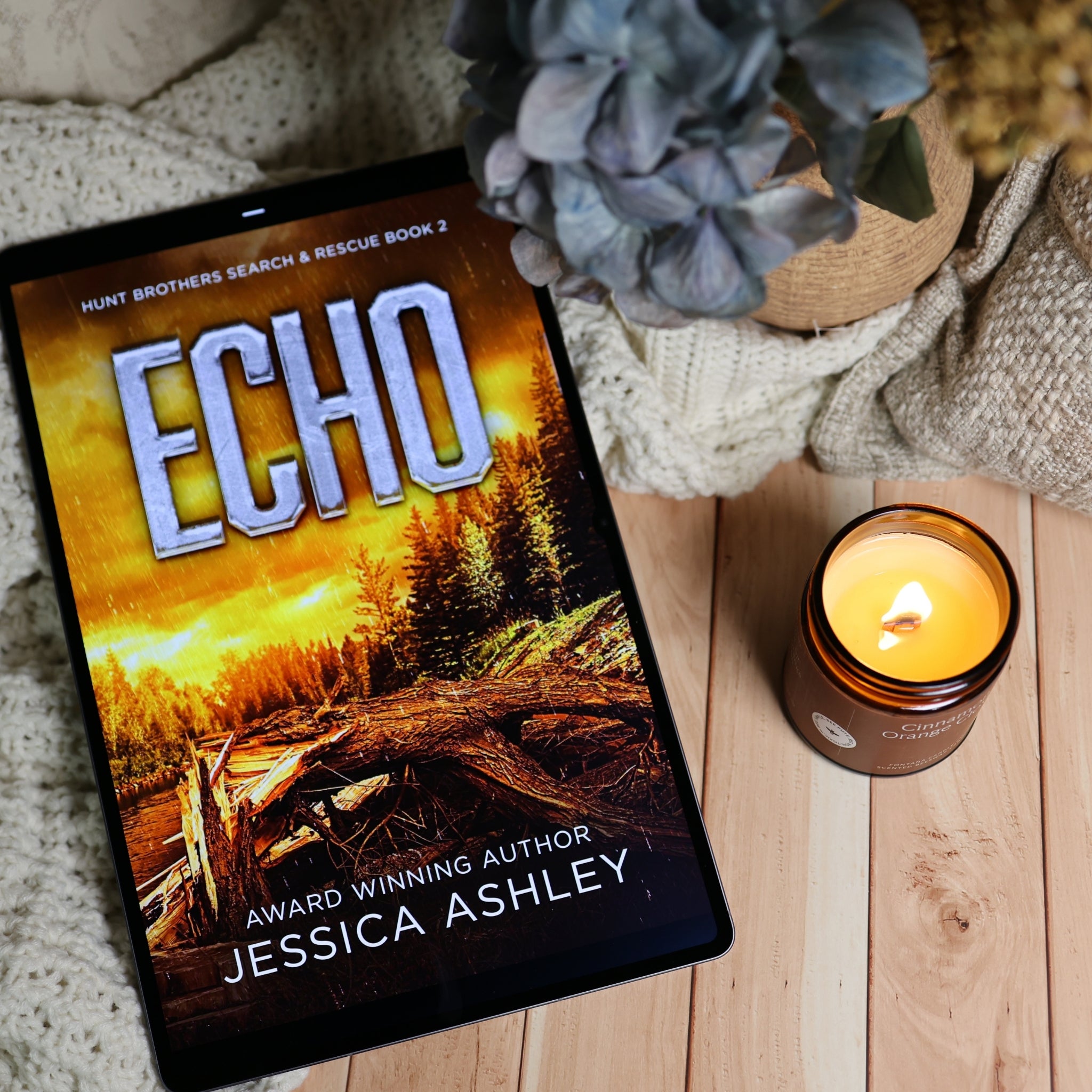 A tablet displaying the book cover of 'Echo' by Jessica Ashley, styled on a wooden surface next to a lit amber candle and a knitted blanket with dried flowers.