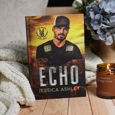 A book titled 'ECHO' by Jessica Ashley is displayed upright on a wooden surface next to a lit candle and a knitted blanket. The book cover features a man in a baseball cap against a warm sunset background.