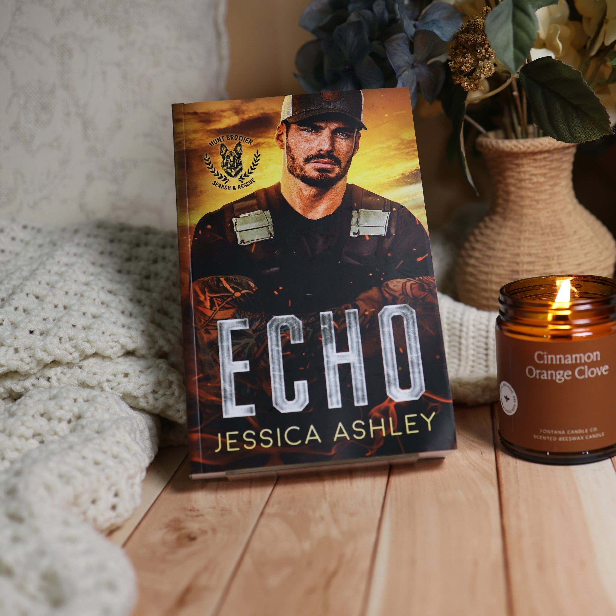 A book titled 'ECHO' by Jessica Ashley stands upright on a wooden surface next to a lit candle in a woven vase with dried flowers and a knitted blanket.