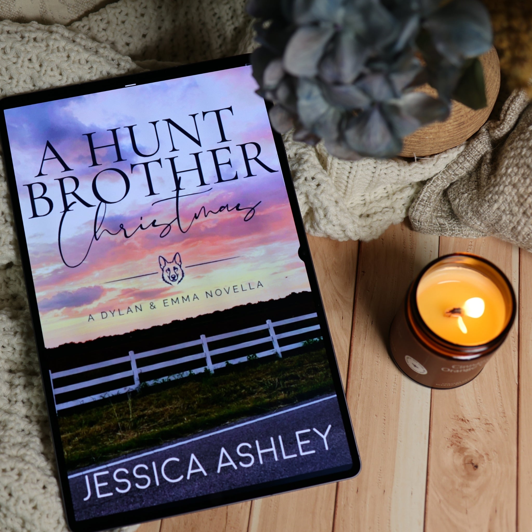 A tablet displaying the book cover of 'A Hunt Brother Christmas' by Jessica Ashley, styled on a wooden surface with a knitted blanket, a lit candle, and a potted plant.