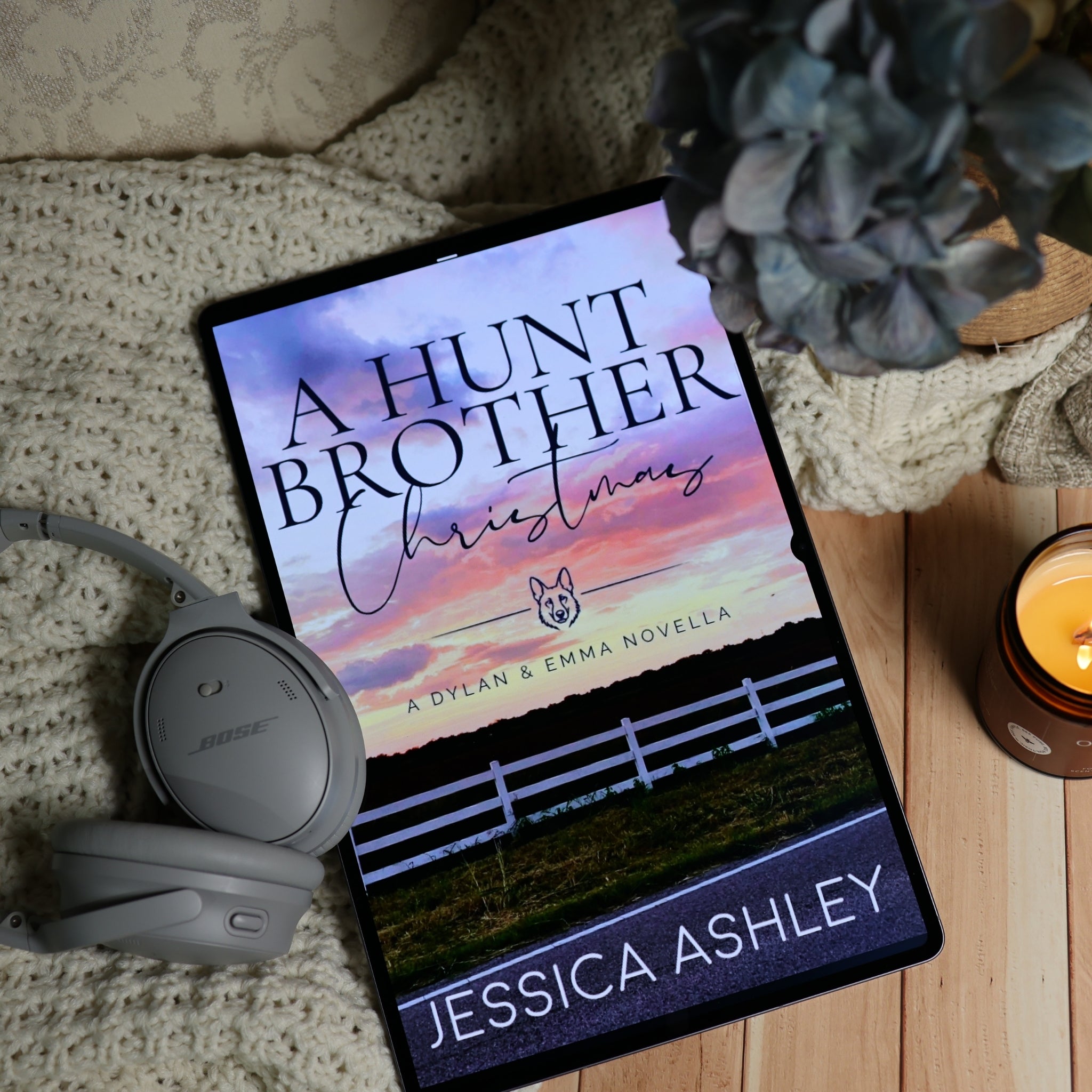 A tablet lying on a knitted blanket on a wooden surface, displaying the book cover for 'A Hunt Brother Christmas' by Jessica Ashley. Gray Bose headphones rest beside the tablet, with a blue flower and a lit candle nearby.