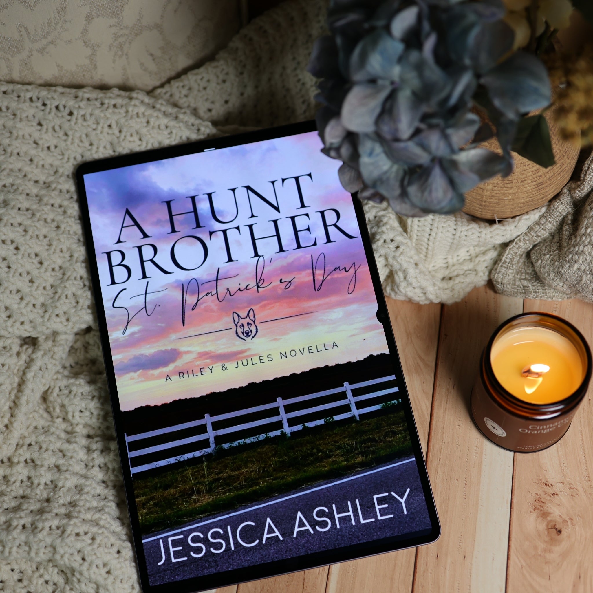 A tablet displaying a book cover for 'A Hunt Brother: St. Patrick's Day' by Jessica Ashley, styled on a wooden surface with a knitted blanket, a lit candle, and a blue flower in a vase.