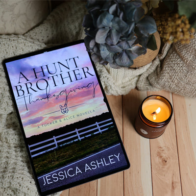 A book cover for the novel 'A Hunt Brother: Thanksgiving' by Jessica Ashley is displayed on a tablet, styled in a cozy flat-lay setting with a lit candle, a knitted blanket, and a potted plant on a wooden surface.