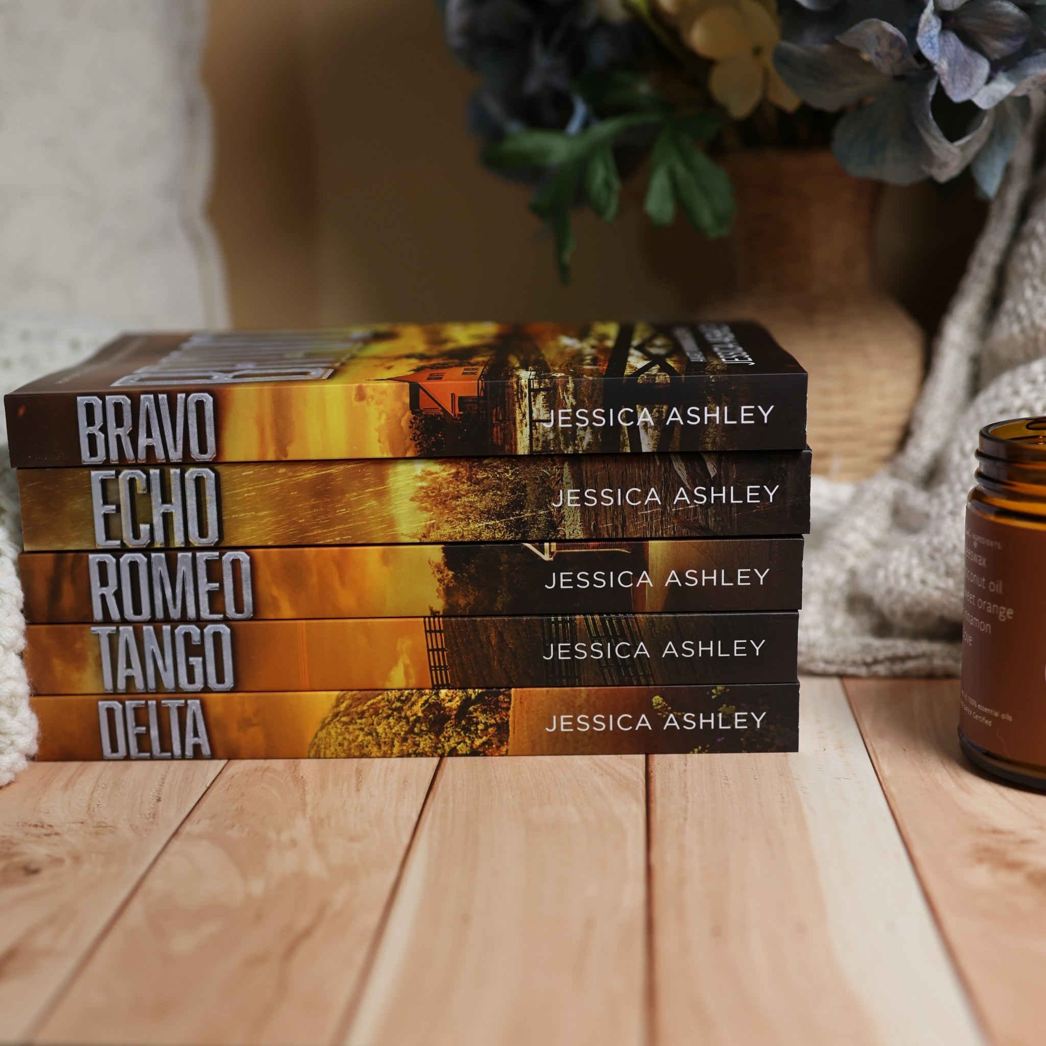 A stack of five romance novels by Jessica Ashley, titled Bravo, Echo, Romeo, Tango, and Delta, displayed on a light wood surface with a cozy background of a knitted blanket and a candle.
