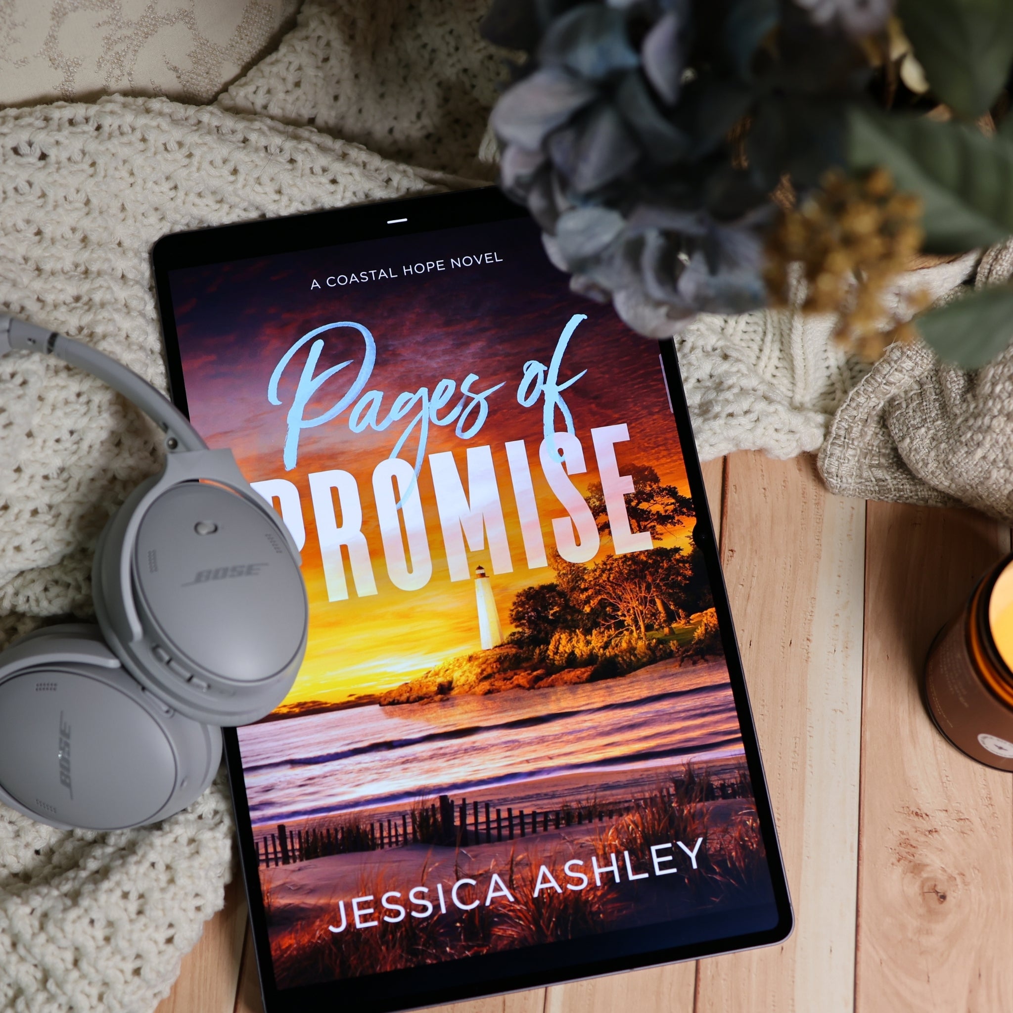 A tablet lying on a wooden surface with a cozy knit blanket, gray Bose headphones, and dried flowers around it. The screen displays the alternate audiobook cover for 'Pages of Promise' by Jessica Ashley.