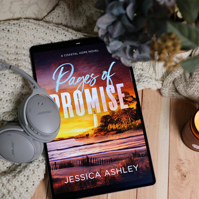 A tablet lying on a wooden surface with a cozy knit blanket, gray Bose headphones, and dried flowers around it. The screen displays the alternate audiobook cover for 'Pages of Promise' by Jessica Ashley.