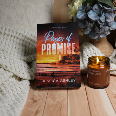 A book titled "Pages of Promise" by Jessica Ashley stands upright on a wooden surface, styled with a cream knit blanket, a lit amber candle, and blue flowers in the background.