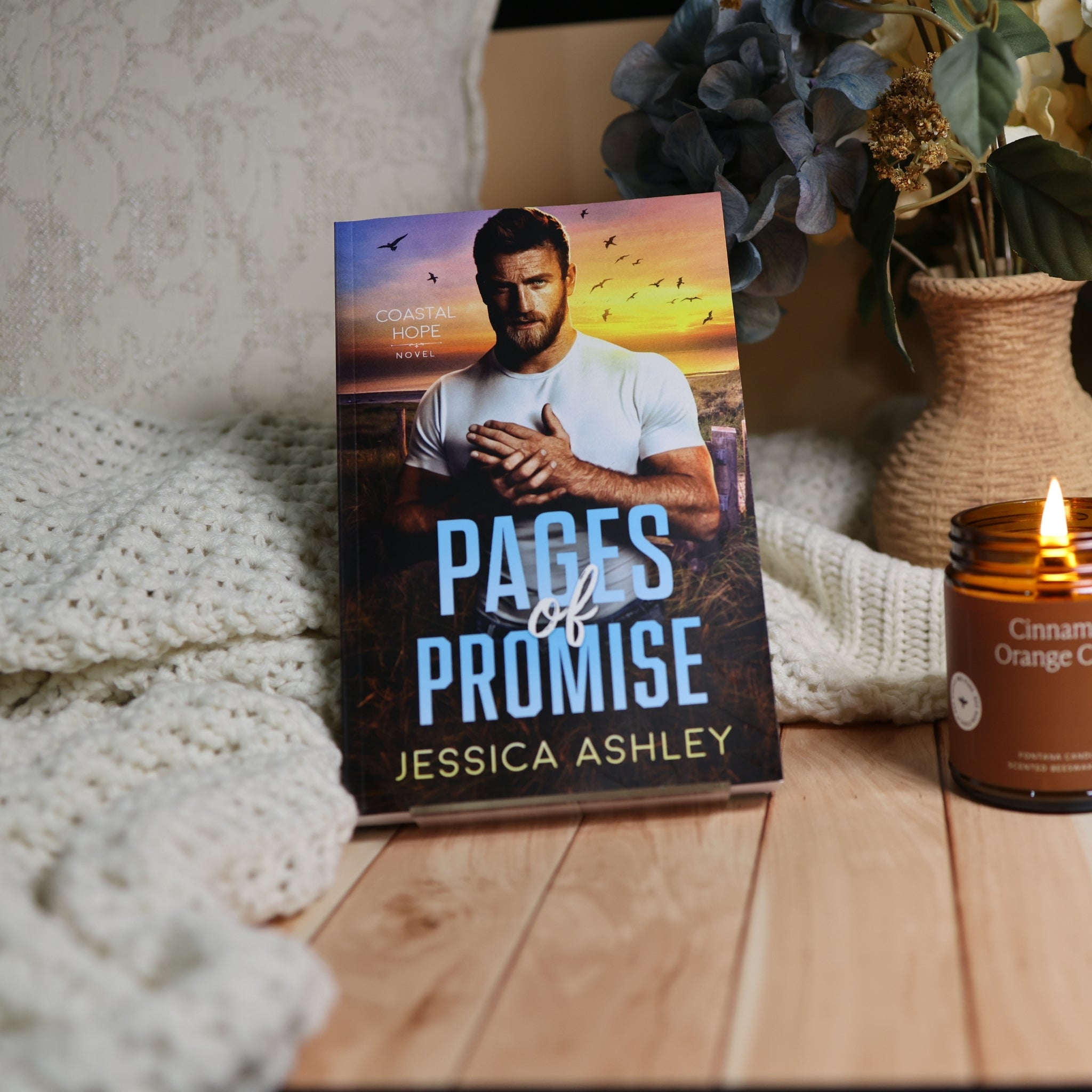 A romance novel book titled 'Pages of Promise' by Jessica Ashley, standing upright on a wooden surface next to a lit candle and a cozy knit blanket.