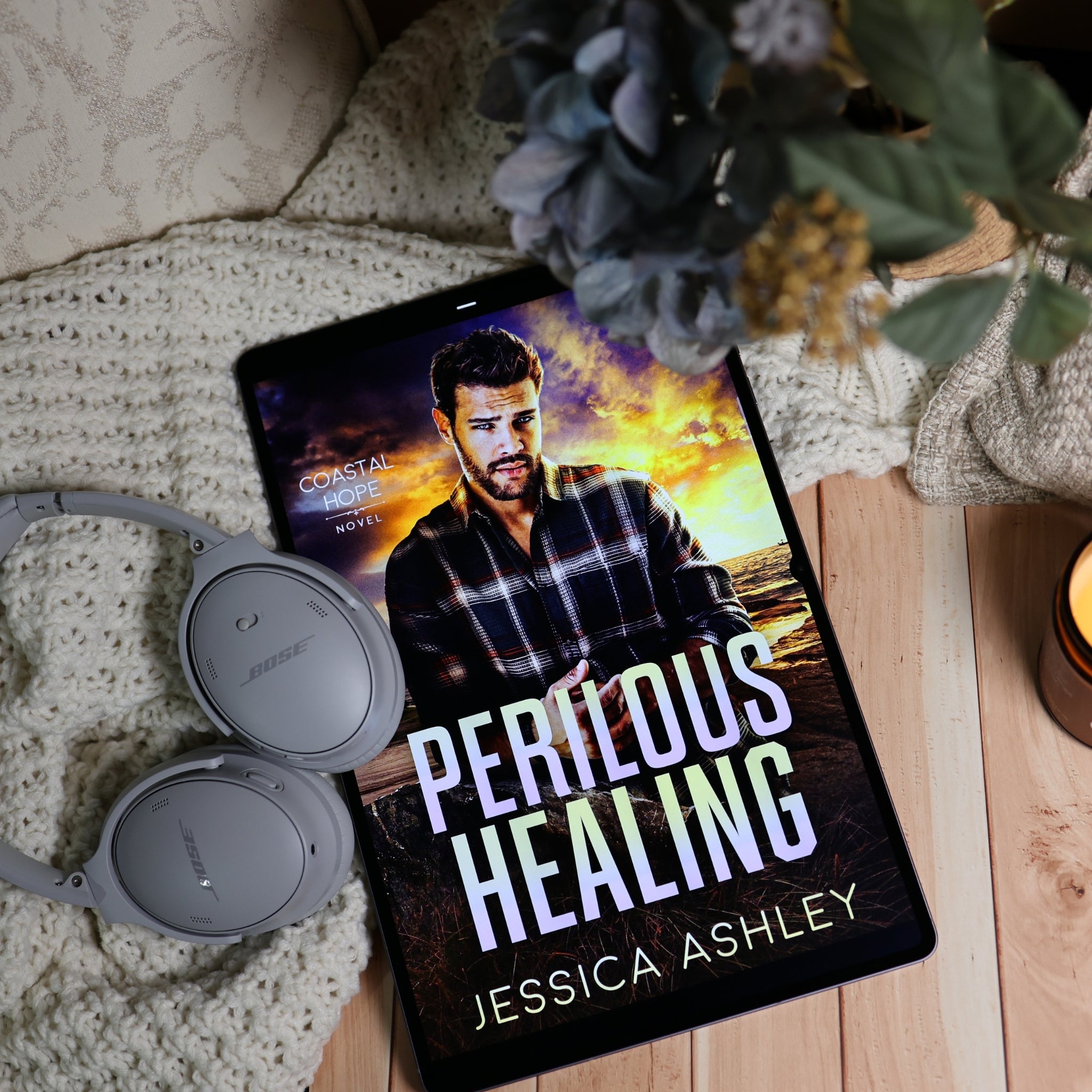 A tablet lying on a light wood surface with a knitted blanket, gray Bose headphones, and dried flowers around it. The tablet screen shows a book cover for 'Perilous Healing' by Jessica Ashley.