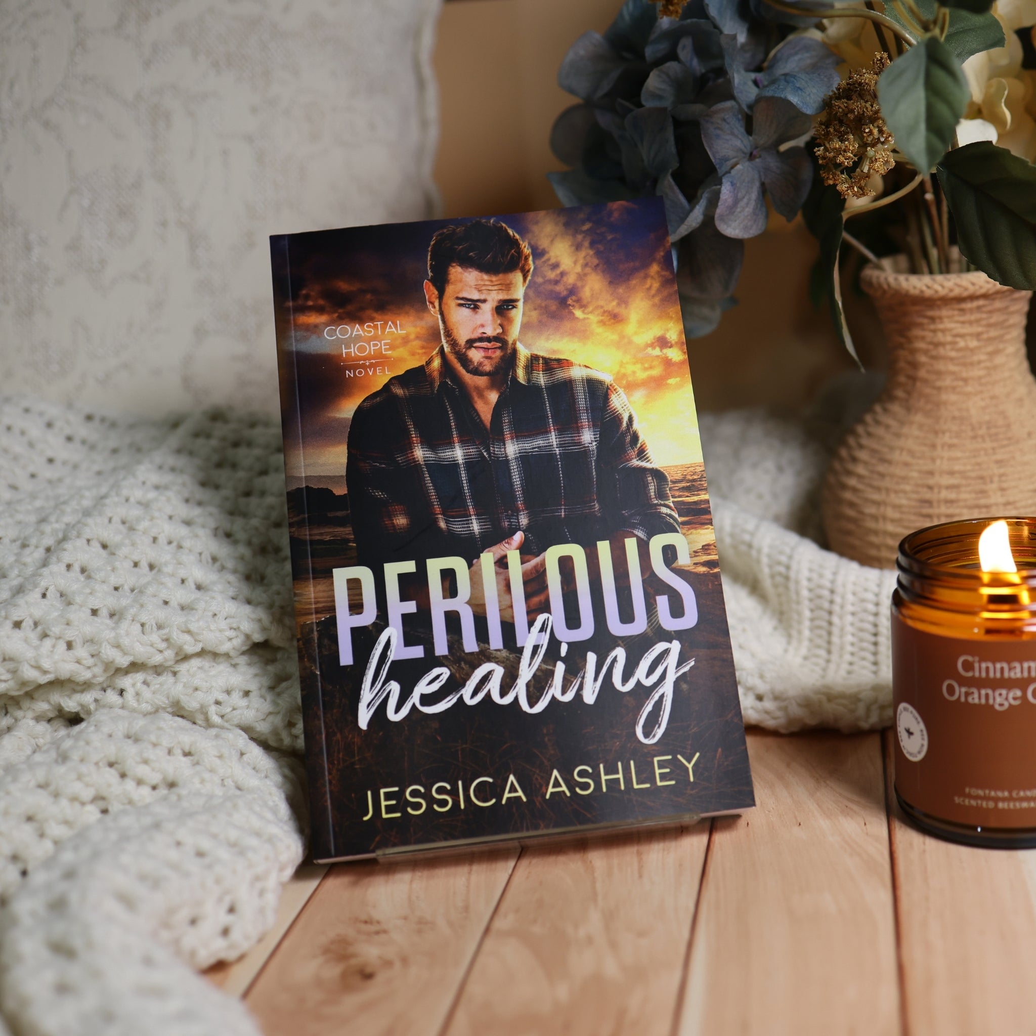 A romance novel book titled 'Perilous Healing' by Jessica Ashley, standing upright on a wooden surface next to a lit candle and a knitted blanket, with a vase of dried flowers in the background.