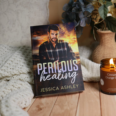 A romance novel book titled 'Perilous Healing' by Jessica Ashley, standing upright on a wooden surface next to a lit candle and a knitted blanket, with a vase of dried flowers in the background.