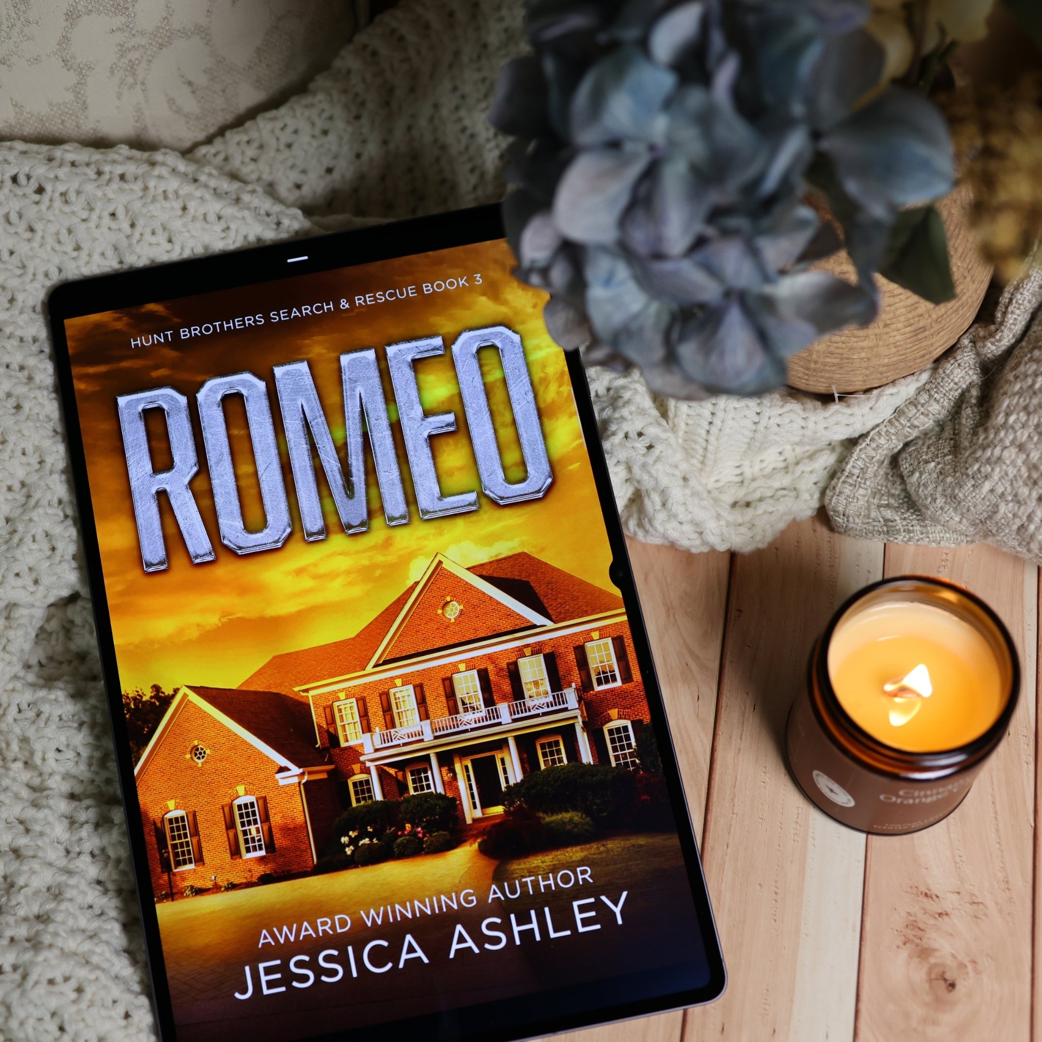 A tablet lying on a wooden surface displays the book cover for 'Romeo' by Jessica Ashley. The scene is styled with a cozy knit blanket, a lit amber candle, and a blue-gray flower in a pot.