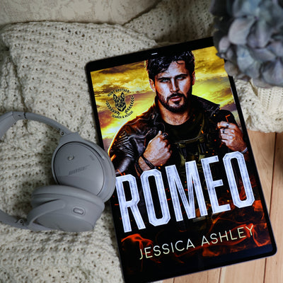 A tablet displaying the book cover for 'Romeo' by Jessica Ashley, styled on a cream knit blanket with gray Bose headphones and a decorative flower in the background.