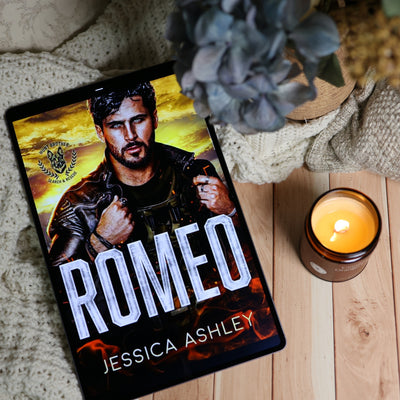 A tablet lying on a wooden surface displays the book cover for 'Romeo' by Jessica Ashley. The scene is styled with a lit candle, a knitted blanket, and a dried flower, creating a cozy atmosphere.