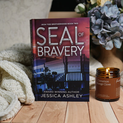 A stack of ten books by Jessica Ashley with dark, dramatic romance novel covers is arranged on a wooden surface next to a lit amber candle. The background features a cozy neutral wall, a soft knit blanket, and a decorative floral arrangement.